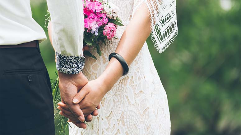 Close up of couple outdoors holding hands. Female wears white wedding dress, holds small bouquet with pink flowers and wears black bracelet. Male wears white dress shirt with black and white pattern cuff and black pants.