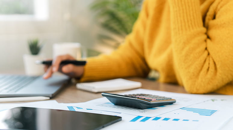 Woman in yellow sweater sitting at a desk looking at her laptop and ipad with her calculator out and graphs on paper.