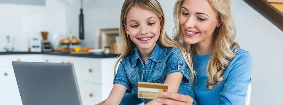 Mother and daughter look at a credit card while working on a laptop on their kitchen counter.