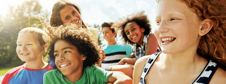 A group of multicultural children sit in a group outdoors, smiling and laughing in the sunshine.