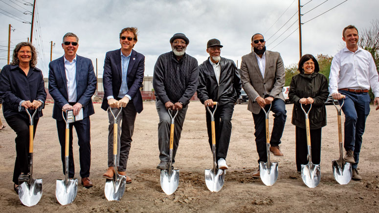 A group of people are lined up holding shovels, their feet held on the heads of the shovels, posing for a photo