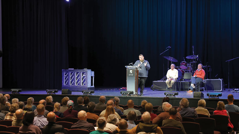 A photo taken inside of an auditorium looking at the stage. A man stands at a podium on the stage talking to the audience, while two other people sit behind him.