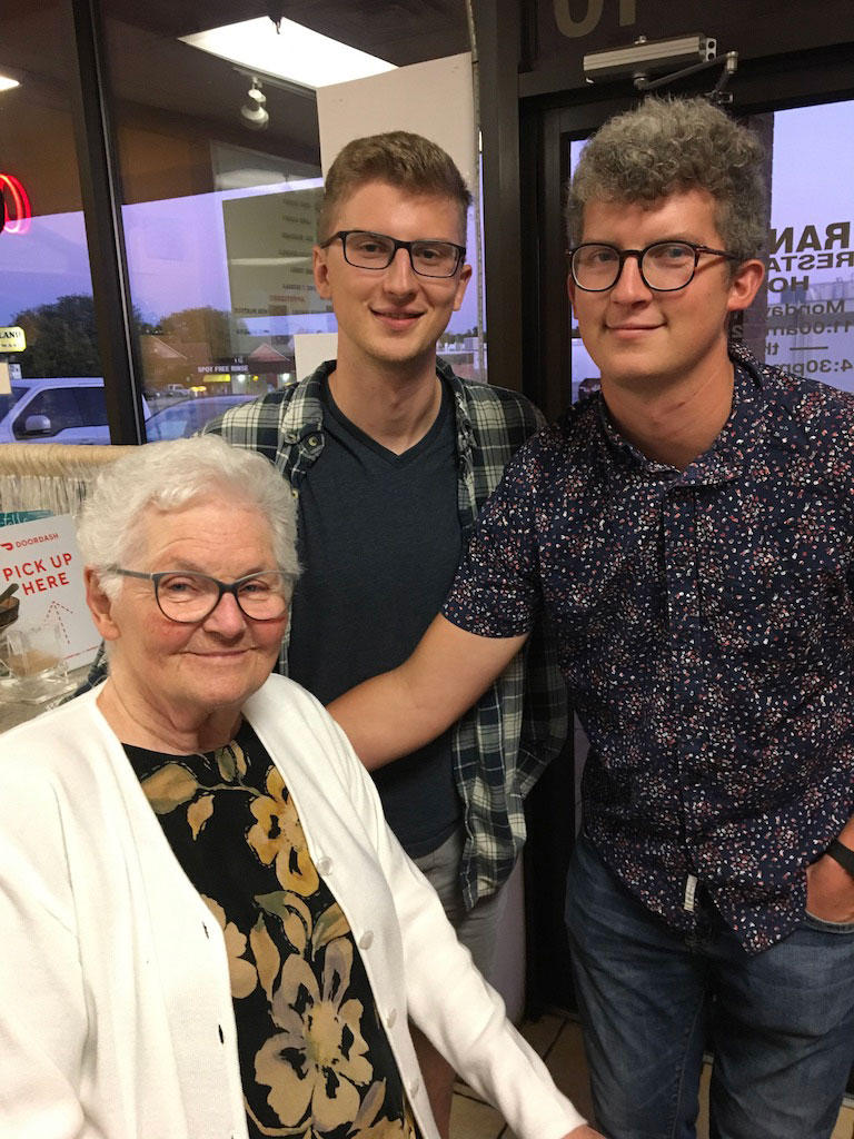 An elderly woman sits, while two younger family members stand beside her, posing for a photo