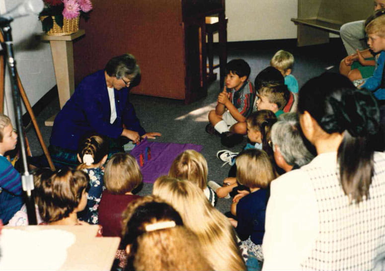 A woman sitting on the floor, reading, while children form a semi-circle around her and watch 
