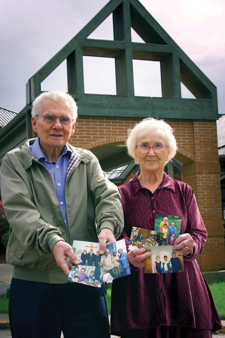 A man and a woman stand in front of a building holding and showing photographs