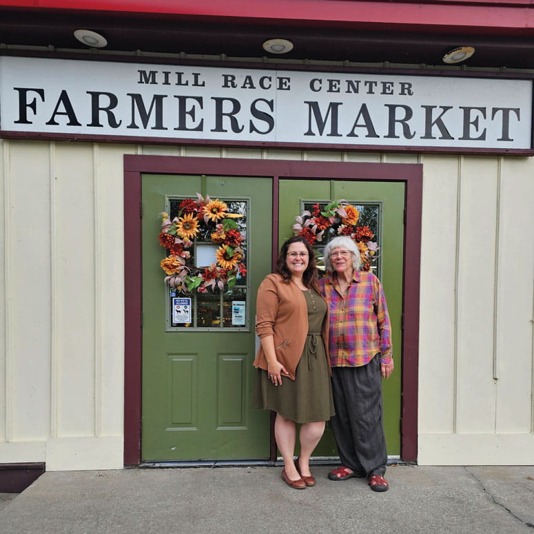 Two woman pose of a photo beneath a sign that reads "Mill Race Center Farmers Market"