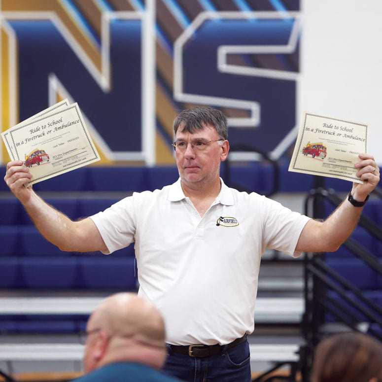 A man looks around at a crowd, holding sheets of paper as if showing them