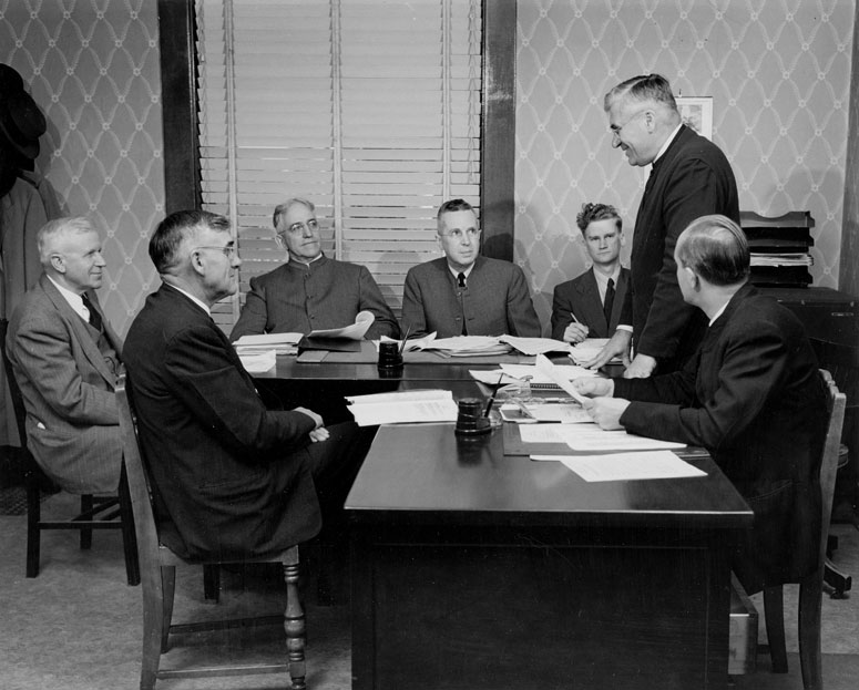 A photo taken in 1950 shows a group of men sitting around two desks in an office room. One of the men is standing, addressing the others. 