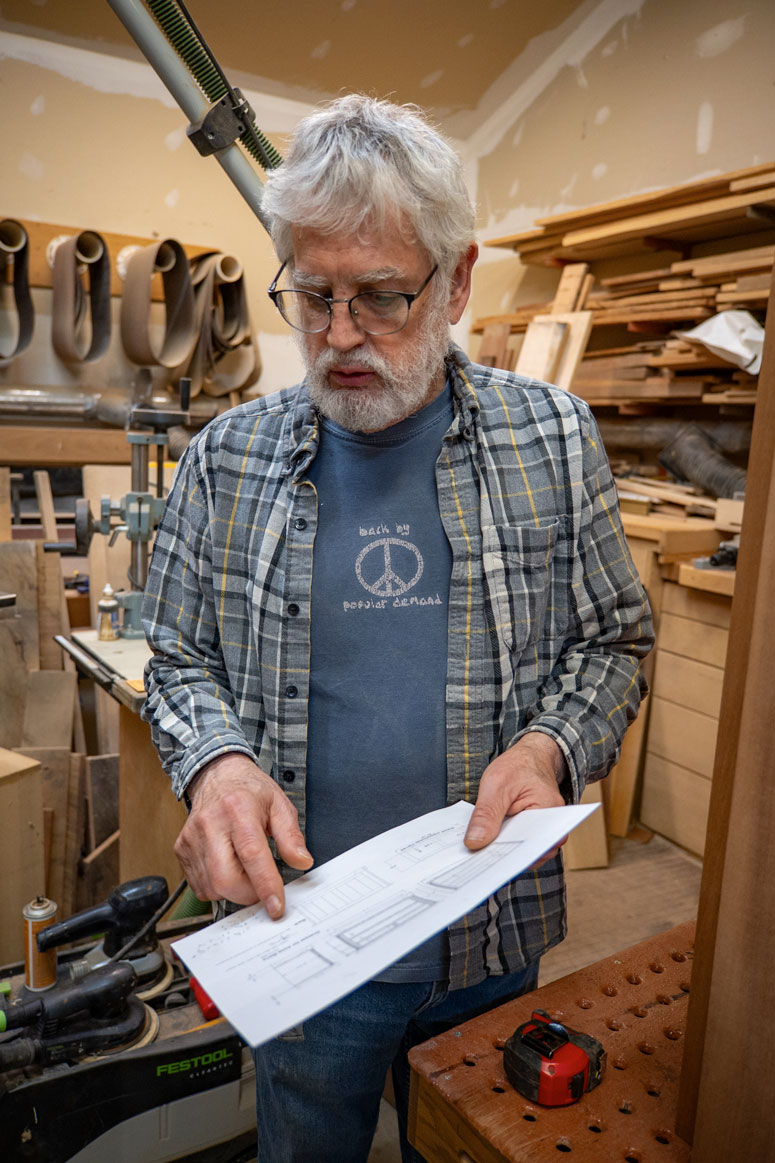 A man stands in his woodworking workshop holding a sheet with measurements for building a wooden dresser