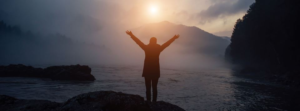 The silhouette of a person standing with their arms outstretched overlooking a body of water with the sun in the background.