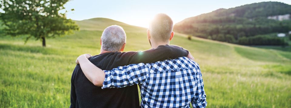 Two men, one older and the other younger have their backs to the camera, looking to the horizon. They have their arm wrapped around the other's shoulder