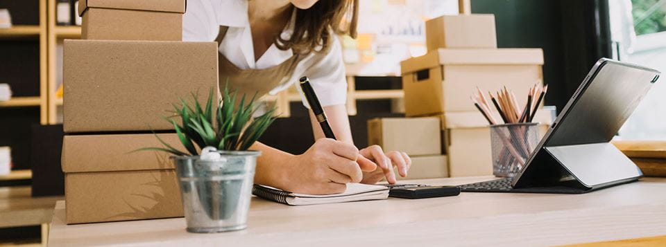 A small-business owner does bookkeeping surrounded by cardboard boxes of inventory.