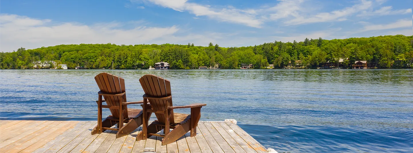 Two adirondak chairs on a dock on a lake with trees and houses on the shore.