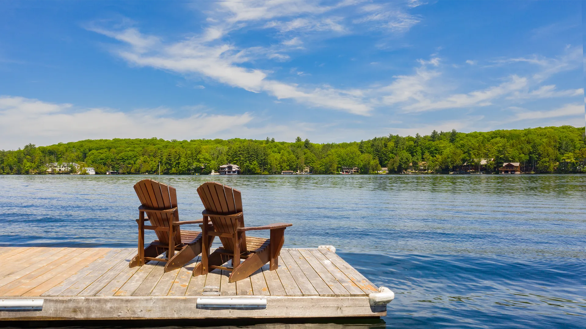 Two adirondak chairs on a dock on a lake with trees and houses on the shore.