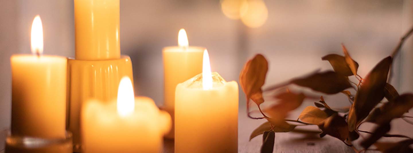 Four lit candles sitting on table cast warm light in dimly lit space with dried orange brown branches decorate the table to the right, creating peaceful, reflective tableau.