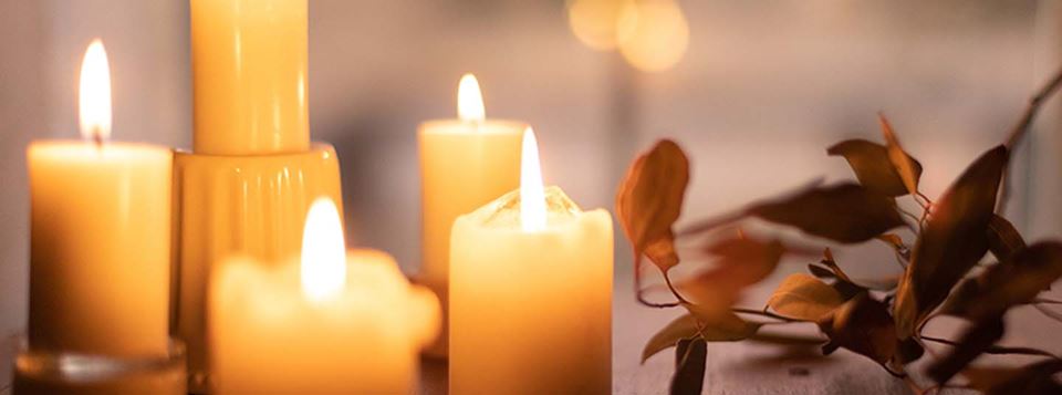 Four lit candles sitting on table cast warm light in dimly lit space with dried orange brown branches decorate the table to the right, creating peaceful, reflective tableau.