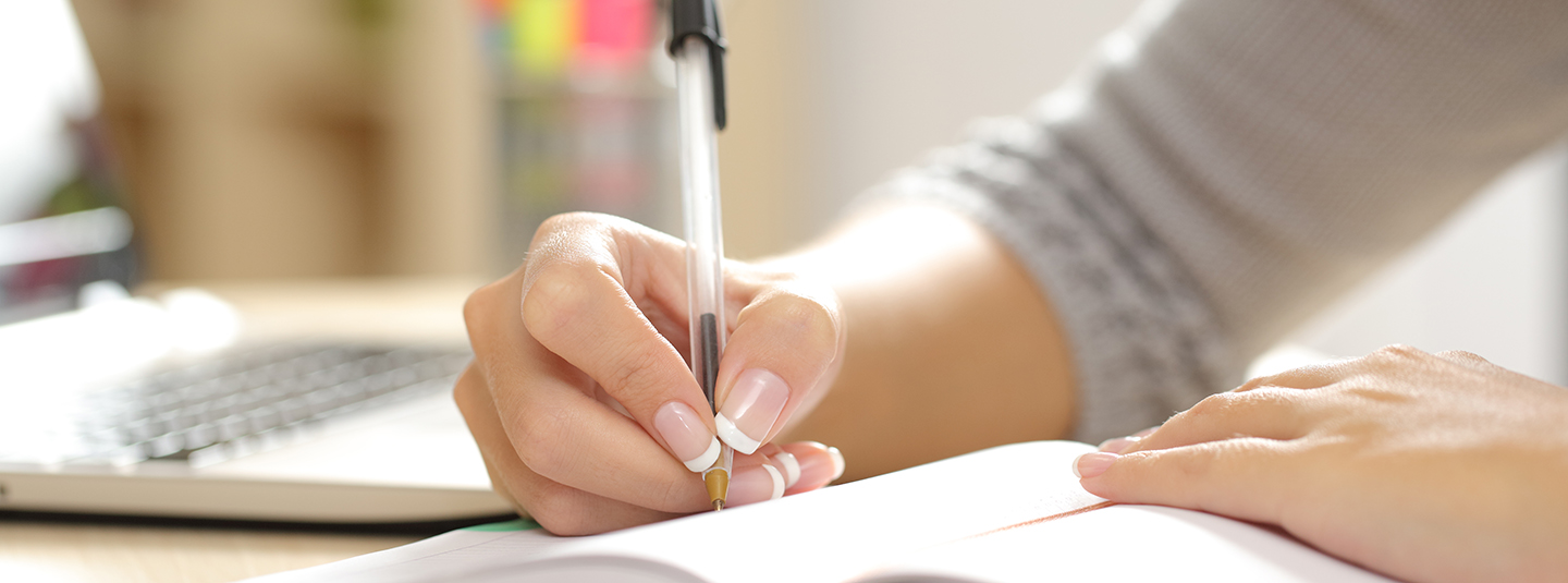 Woman writing in notebook with pen and laptop to the side