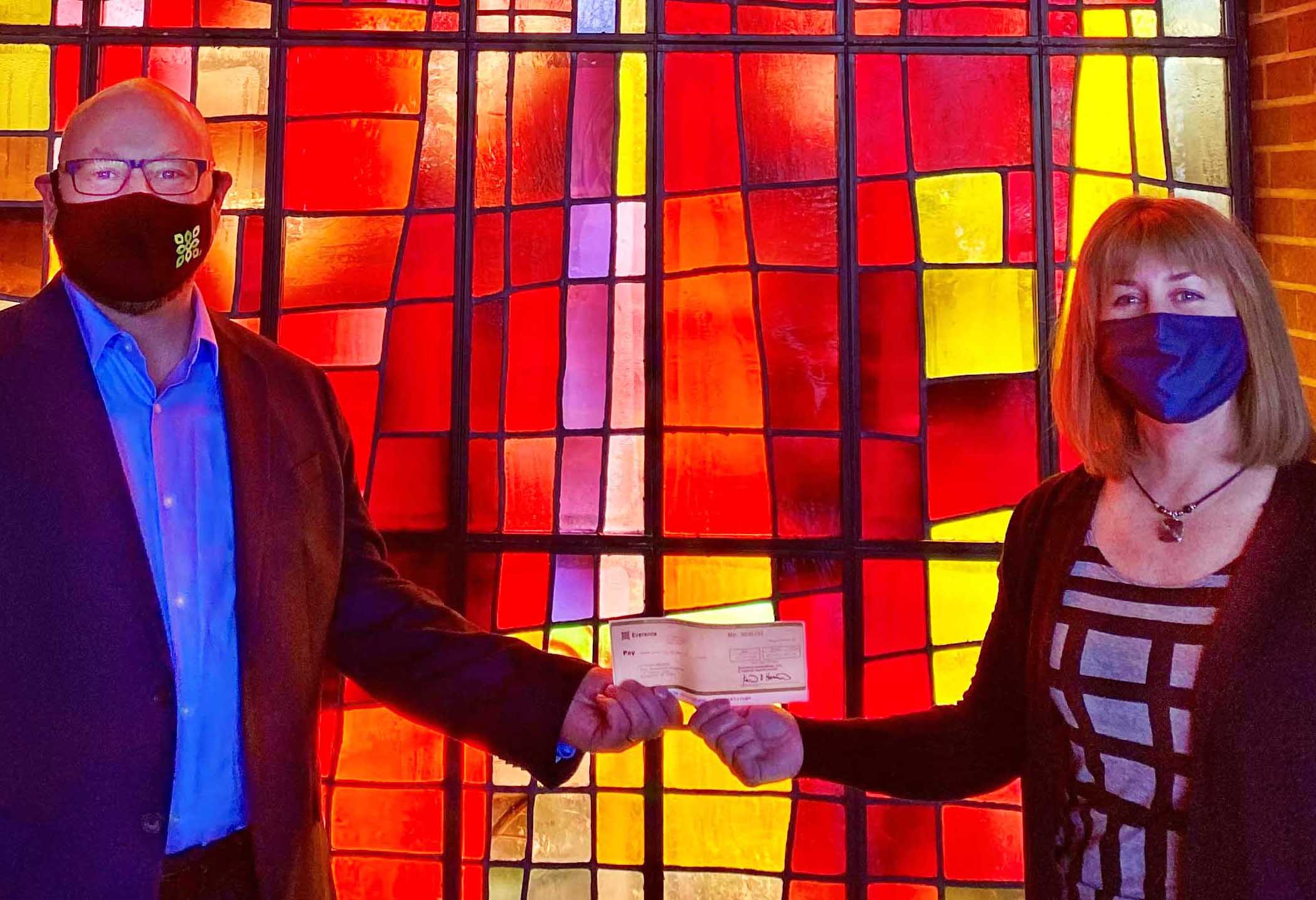 Everence Stewardship Consultant Randy Nyce with Everence mask presents a check to Donna Halteman with blue mask, founder and coordinator of the Bean Bag Food Program in front of stained glass window with red 