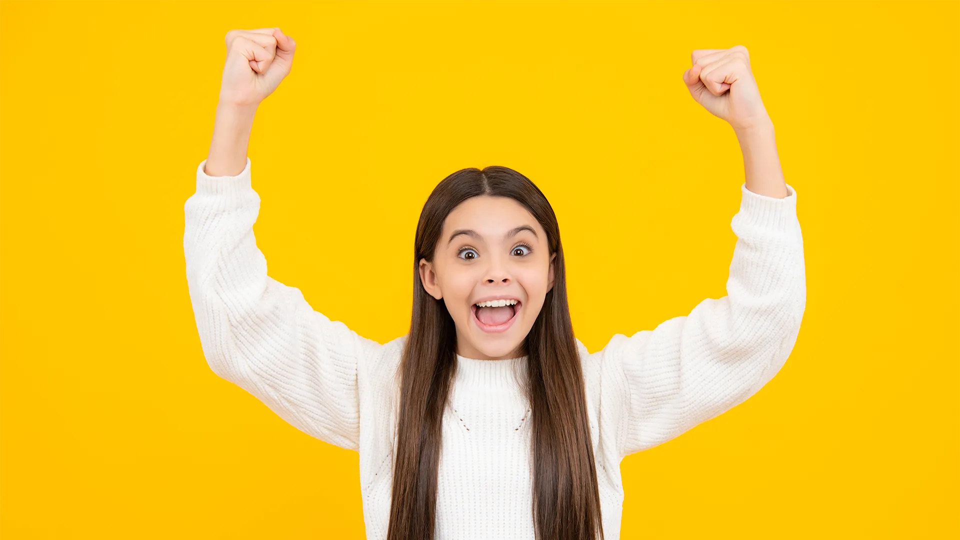 Preteen girl with dark hair raising both fists in the air with enthusiasm and excitement like she won in front of a yellow background.