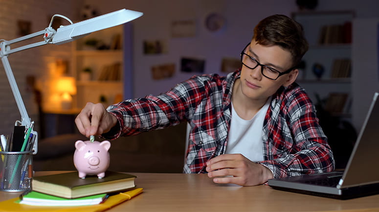 Teen putting money in his piggy bank at a desk