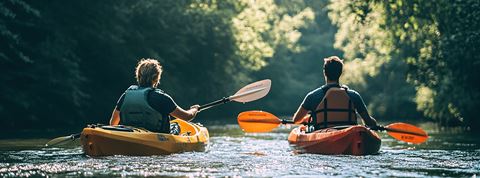 Two kayakers paddle down away down the river surrounded by trees