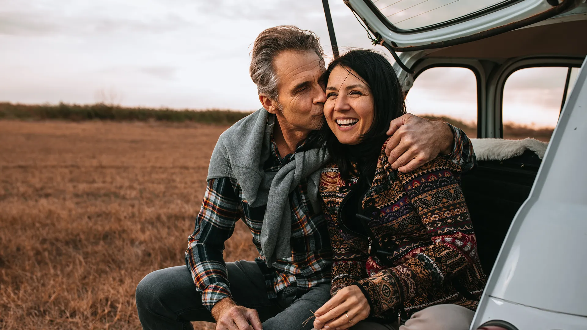 Middle-aged couple with sweaters sit on the back of a car looking at a fall scene.