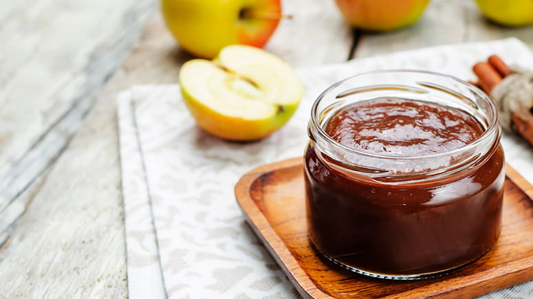 Apple butter in a jar sitting on a marble counter surrounded by apples