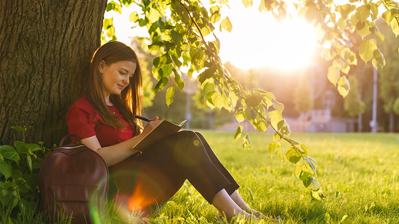 girl in red shirt sitting under a tree writing in a brown notebook
