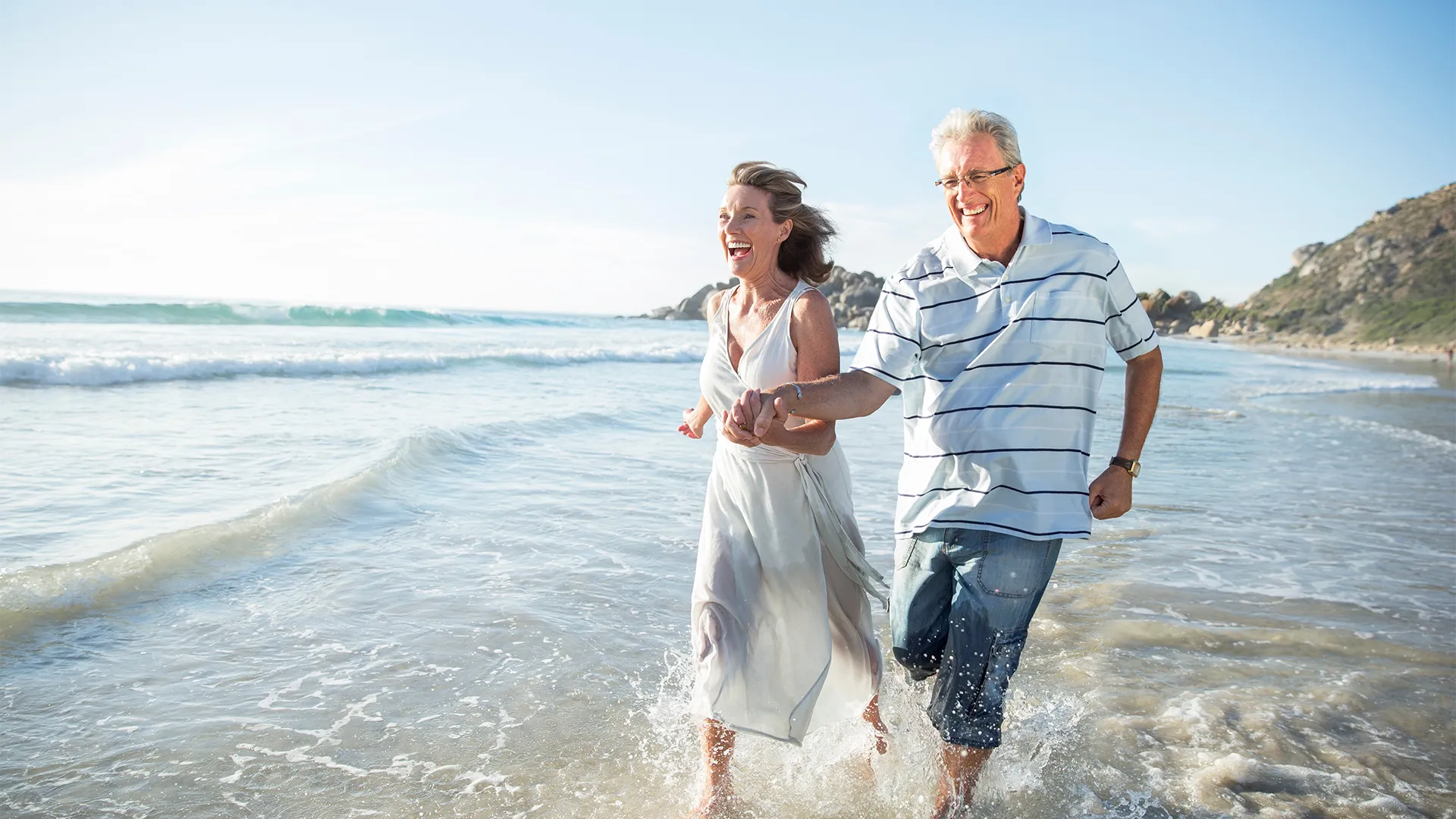Older couple runs into the water at the beach while smiling.