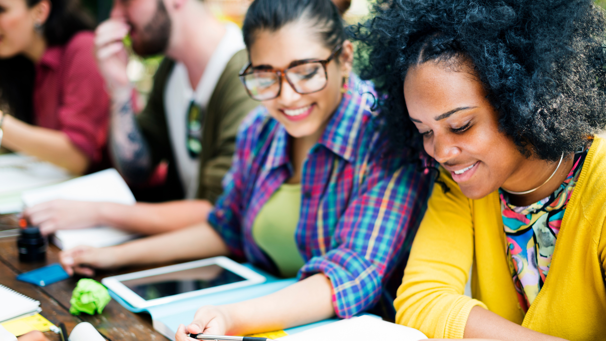 A young Latina woman in a blue plaid shirt and young African American woman in a yellow cardigan study with friends.