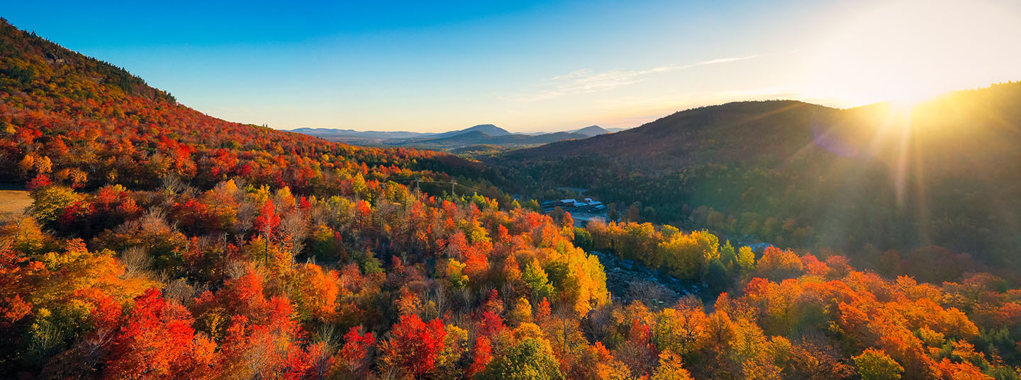 The sun peaks out over a mountain range covered in colorful fall trees.
