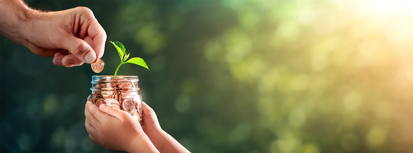 One hand puts a penny in a jar full of pennies with a small plant growing out of it being held by another pair of hands with a leafy and sunny background.