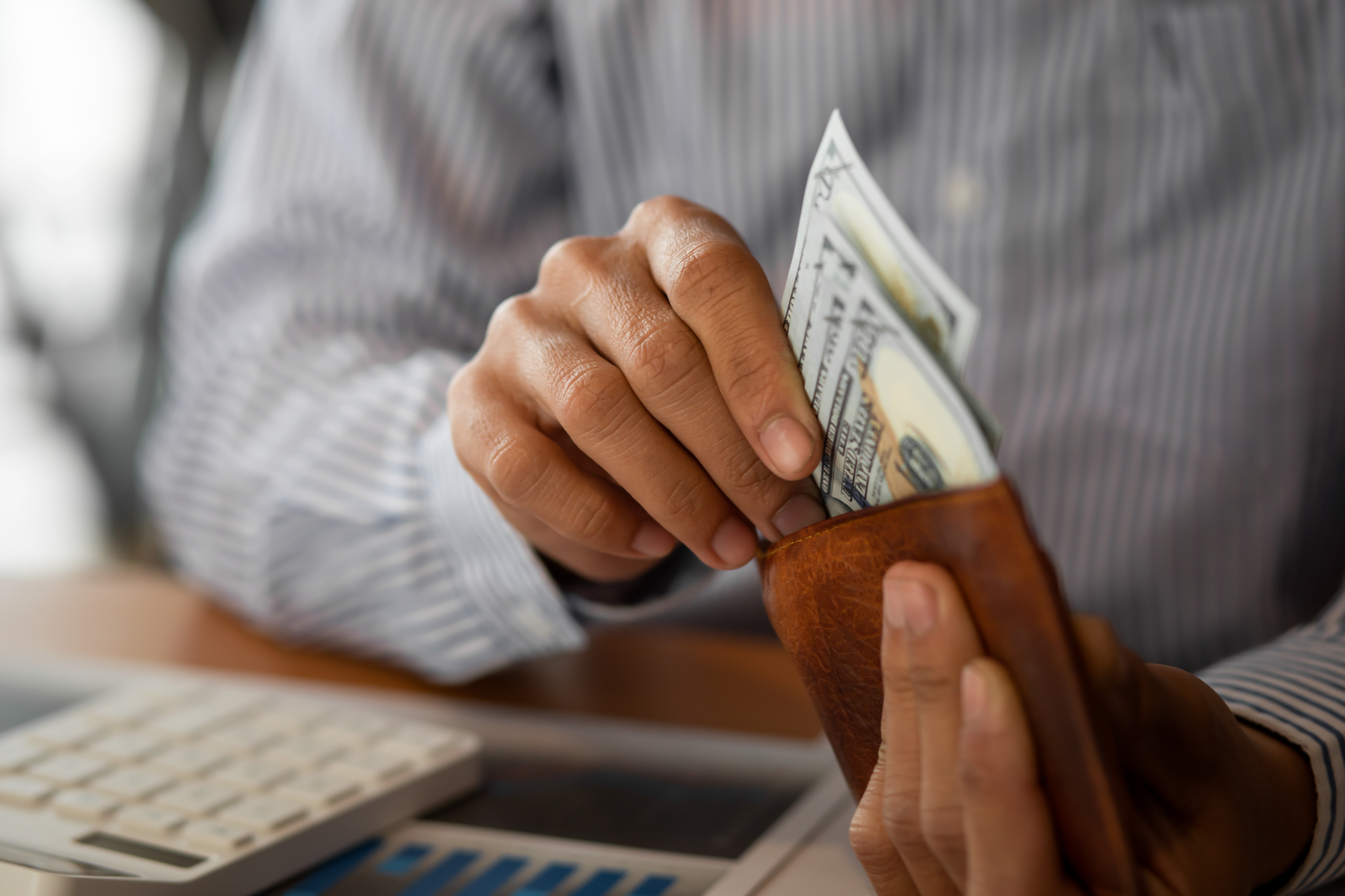 A man puts money in his wallet while sitting over a calculator and a desk.