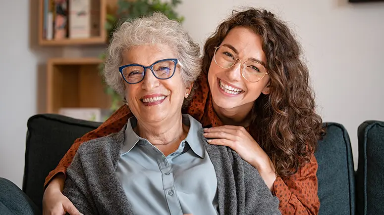 Grandmother smiling with young adult granddaughter both sitting on a couch.